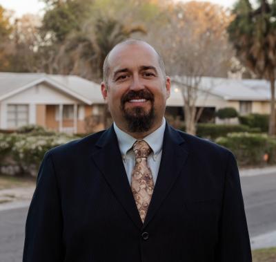 A person wearing a dark suit jacket, light dress shirt, and patterned tie is standing outdoors in a residential neighborhood.