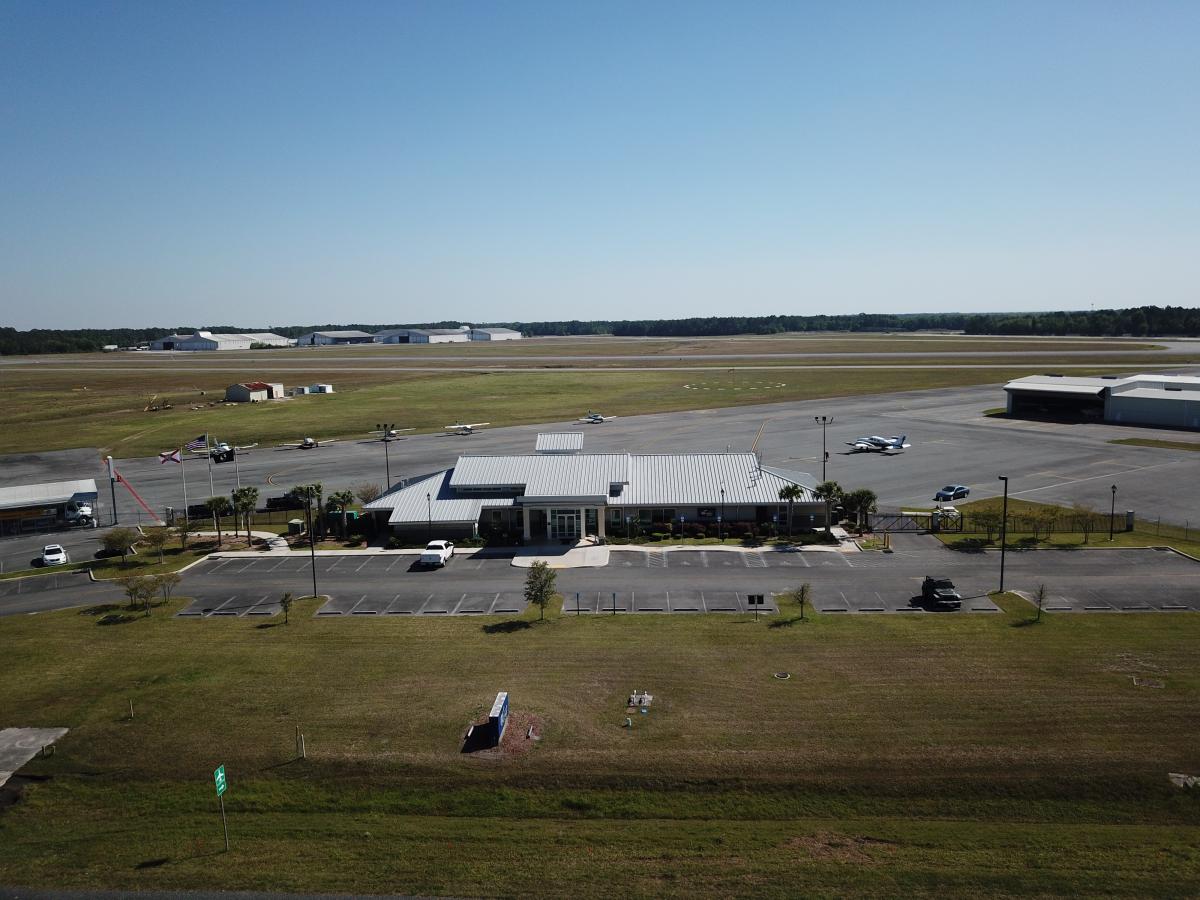 The airport terminal office building, five small airplanes parked behind it and two to the right of the building, runway behind.